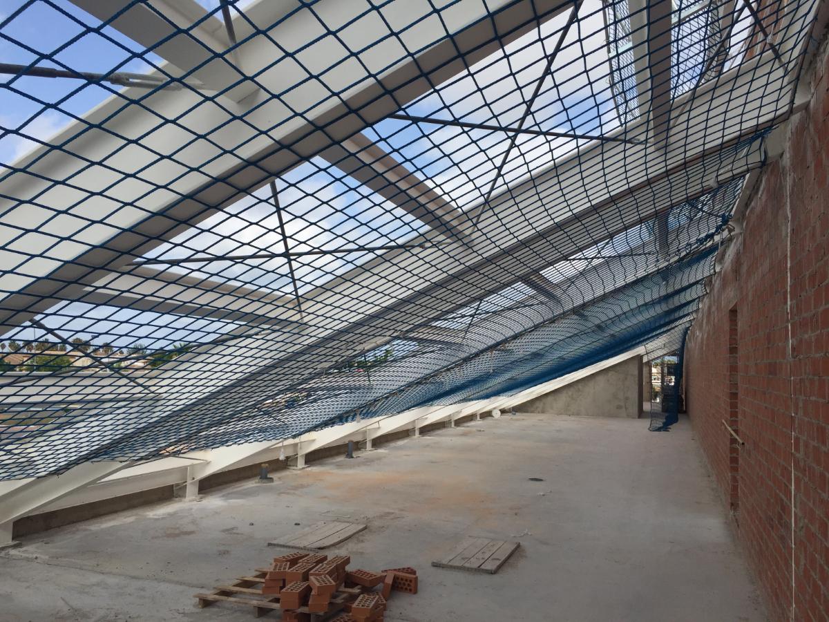 Red de seguridad horizontal (Tipo S o U) de color azul y malla cuadrada instalada bajo la estructura metálica del techo en una planta de un edificio en construcción, con paredes de ladrillo y suelo de hormigón, para protección contra caídas.